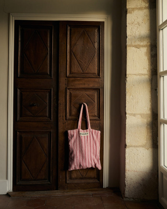 Rosa Tote Bag mit roten Streifen hängt am Griff einer alten dunklen Holztür neben einer hellen Steinwand
