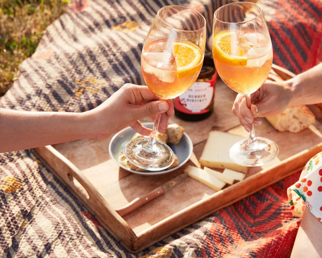 Anstoßen mit zwei Gläsern Hibiskus Spritz mit Orangenscheibe beim Picknick, Holztablett mit Käse