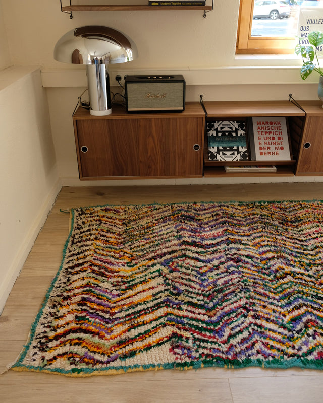 Bunter Zickzack-Teppich von der Schmalseite aus gesehen, dahinter ein Sideboard aus Holz