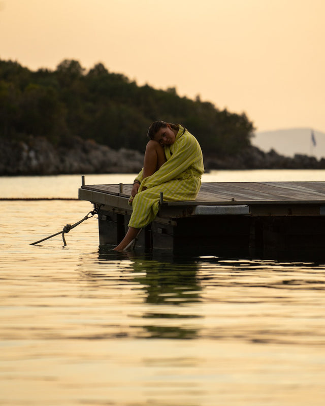Frau im Bademantel NARAM Pristine & Neon Yellow sitzt im Abendlicht auf einem Badesteg am Meer