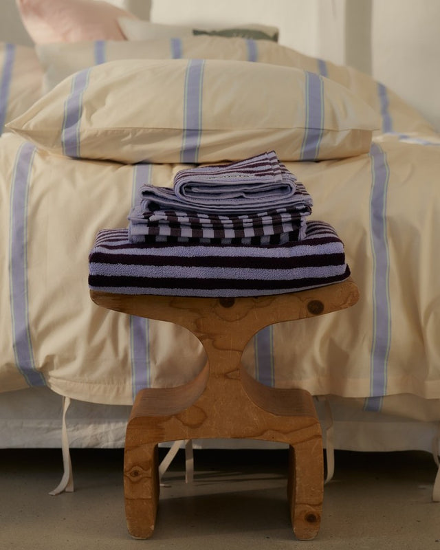 Gefaltete NARAM-Handtücher in Weinrot und Lavendelblau gestapelt auf einem Holzhocker vor dem Bett