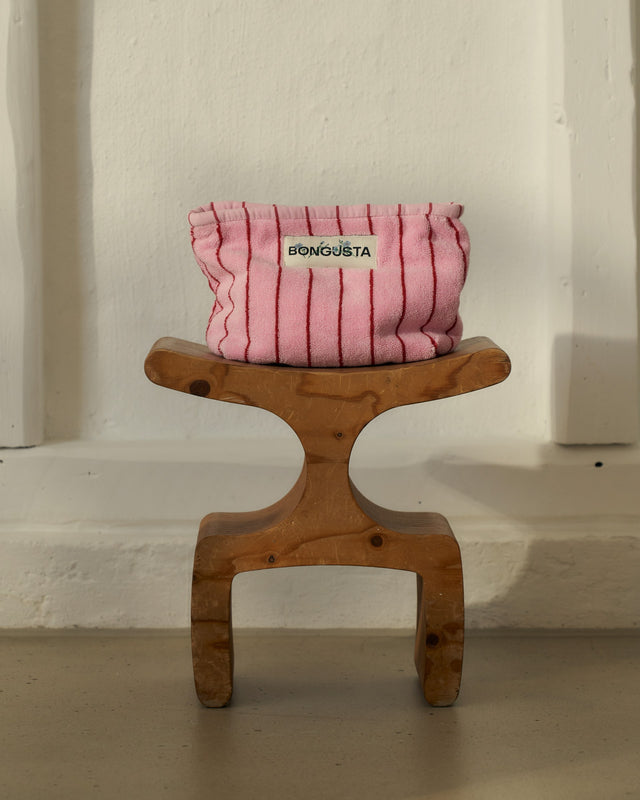 Rosa gestreifter Kulturbeutel NARAM auf einem geschwungenen Holzhocker vor einer weißen Wand