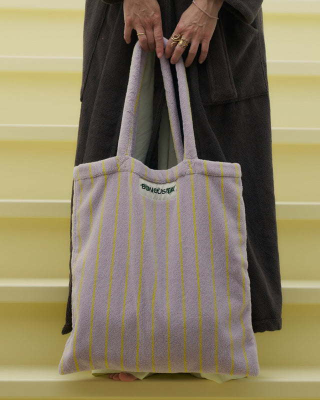 Fliederfarbene Tote Bag mit gelben Streifen, mit beiden Händen vor einem dunklen Bademantel gehalten, auf einer hellgelben Treppe