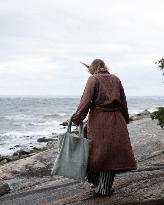 Frau im Bademantel geht über Felsen am Meer und trägt die salbeigrüne Tote Bag NARAM in der Hand