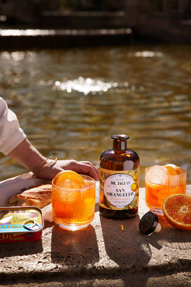 Zwei Gläser San Orangello mit Eis und Orangenscheibe, dazwischen die Flasche, daneben Baguette und eine Dose Sardinen