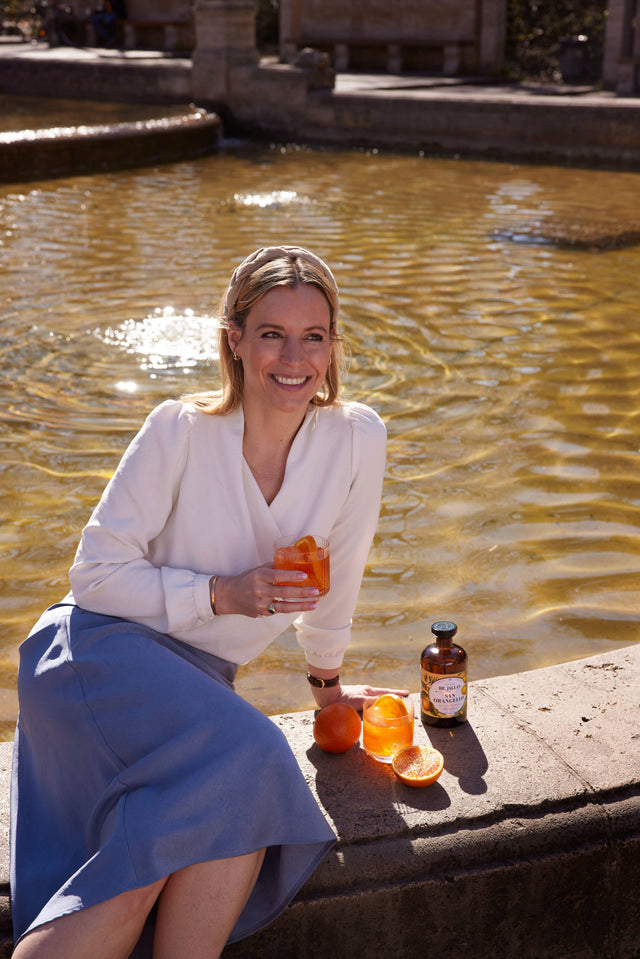 Frau in weißer Bluse sitzt mit einem Glas San Orangello am Brunnenrand, neben ihr Flasche und Orangen