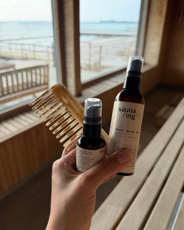 Hand hält den Sauna Serum Mist und eine kleinere sauna/ing-Flasche in einer Sauna mit Blick aufs Meer, daneben ein Holzkamm