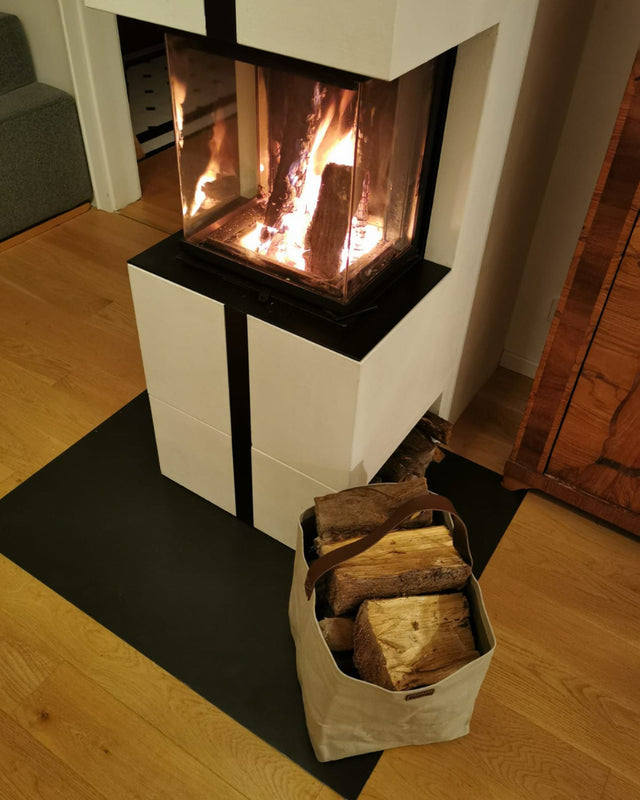 Hellgraue Tasche mit Kaminholz gefüllt neben einem weißen Kamin, in dem ein Feuer lodert.